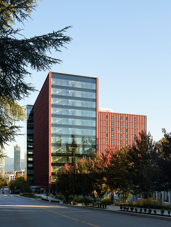 Modern glass building with red accents surrounded by trees and a city backdrop. 